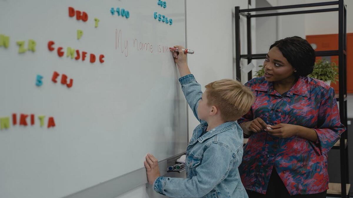 Learner writing on board as the teacher instructs him